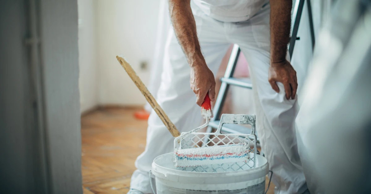 A painter in white coveralls loads a paint roller from a bucket, with a wooden paint stick and a ladder nearby.