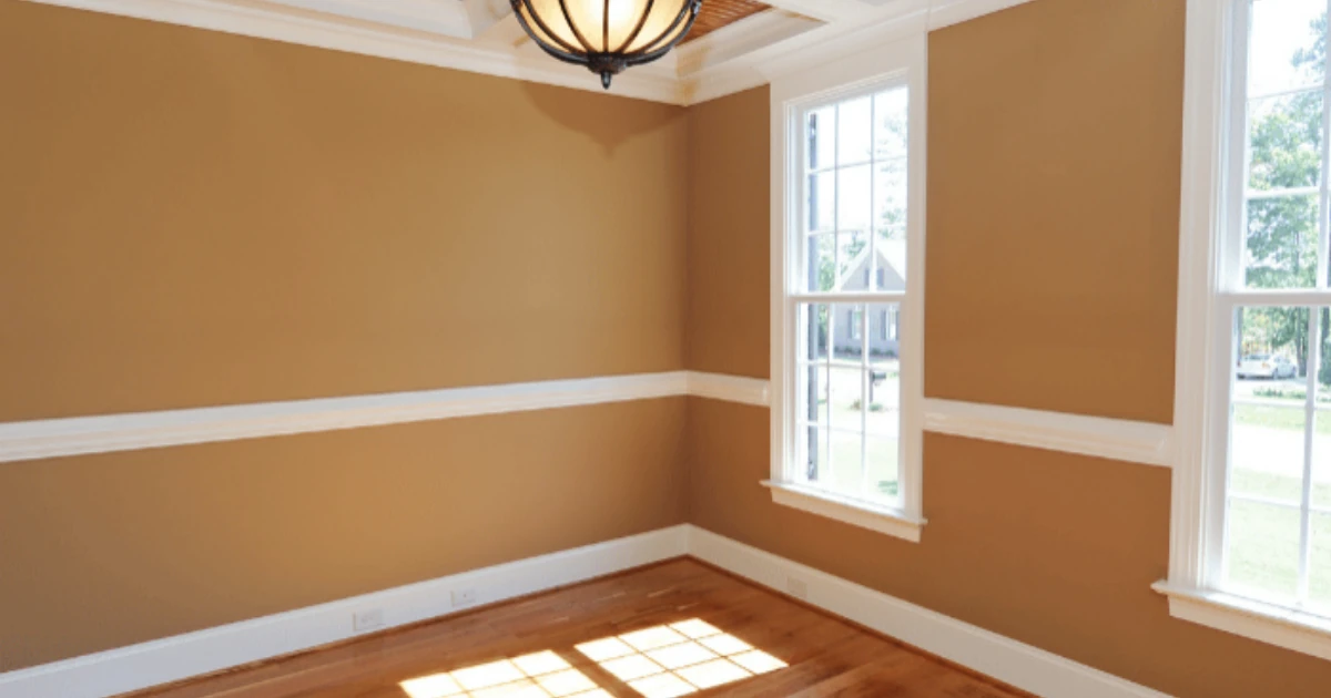 Empty dining room with mustard-gold painted walls, a white chair rail and crown molding, a hanging ceiling light fixture, two large windows, and hardwood flooring.