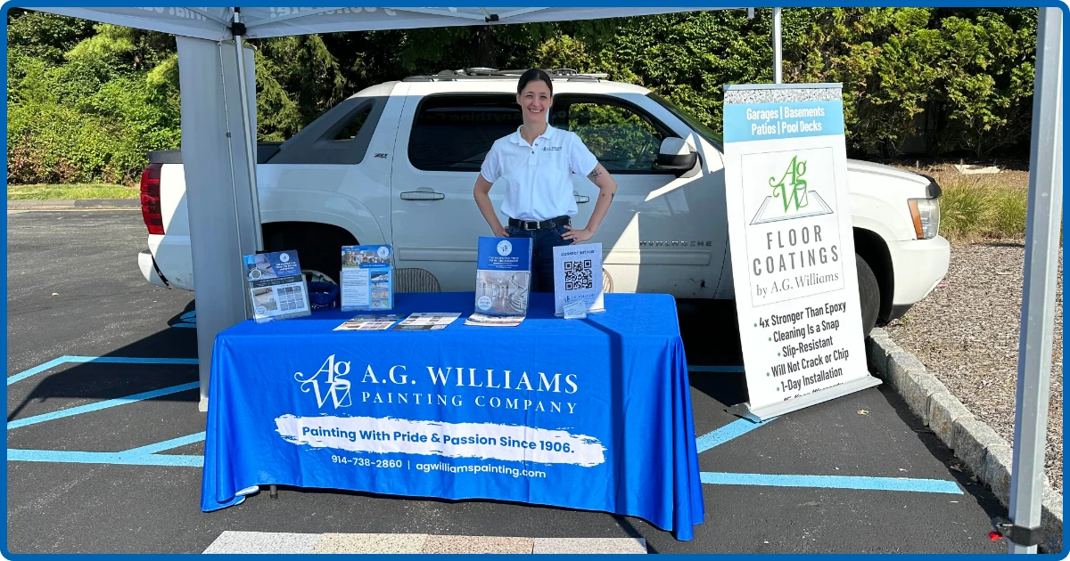 A woman in a white A.G. Williams Painting Company polo shirt stands behind a blue-skirted table with a banner reading "Painting With Pride & Passion Since 1906," under a canopy tent with a floor coatings sign and a white SUV parked behind her.