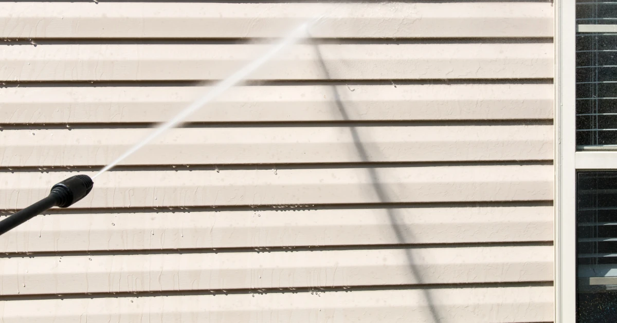 A pressure washer nozzle sprays a jet of water across beige horizontal vinyl siding beside a window.