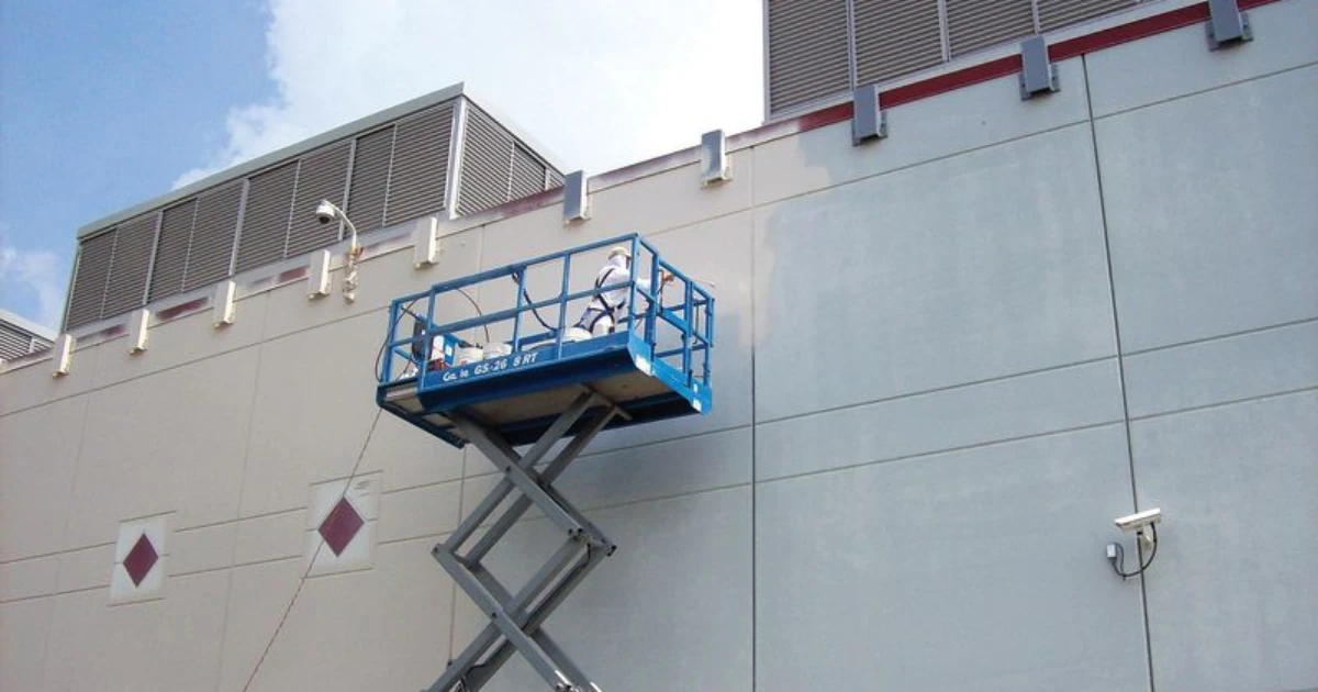 A worker in white coveralls stands in a blue scissor lift painting the exterior wall of a large commercial building.