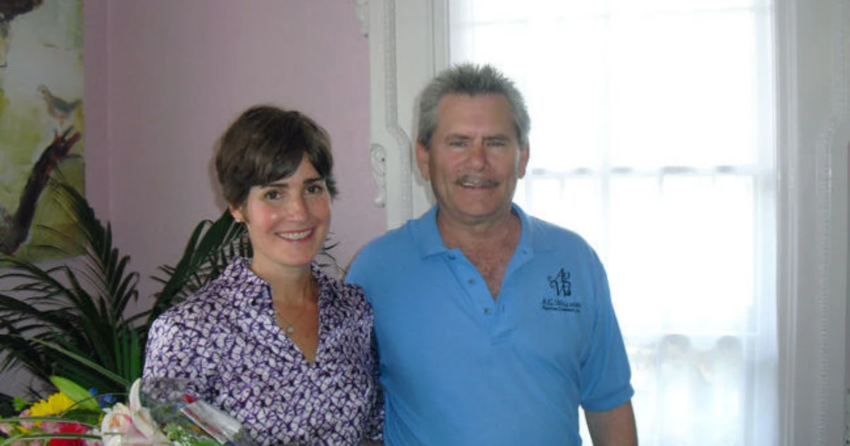 A man in a light blue A.G. Williams polo shirt stands with a woman indoors near pink walls with white molding and a floral painting.