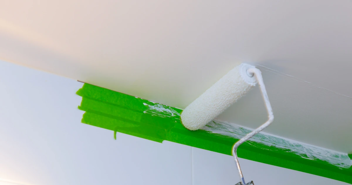 A close-up of a paint roller applying white paint along a ceiling edge protected by green painter's tape.