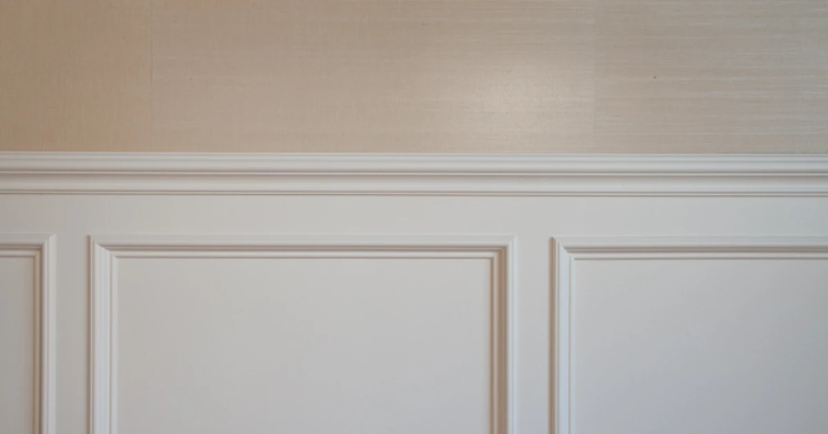 Close-up of white painted wainscoting panels below a chair rail, with tan grasscloth wallpaper above.