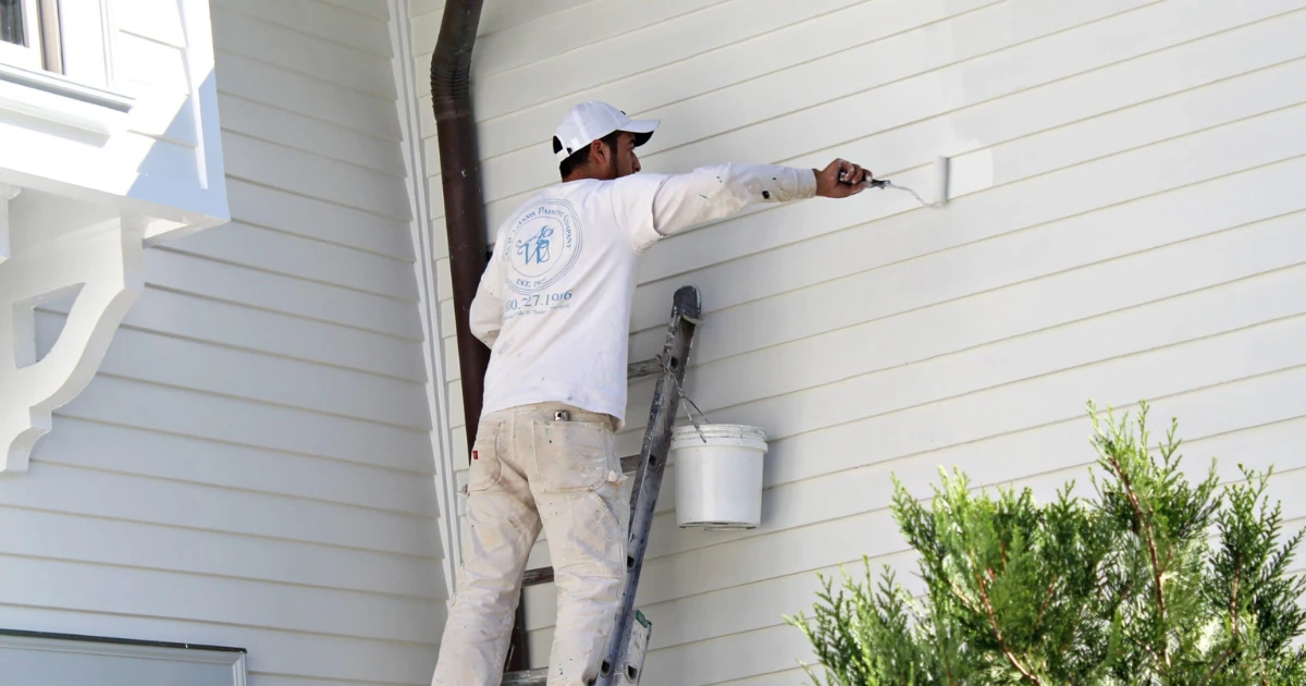 A painter in an A.G. Williams Painting Company shirt standing on a ladder, using a paint sprayer on light-colored vinyl siding.