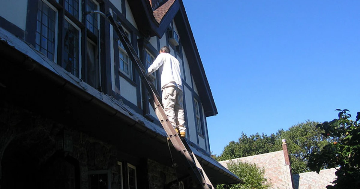A painter stands on a tall extension ladder against a Tudor-style house, painting trim near the roofline under a clear blue sky.