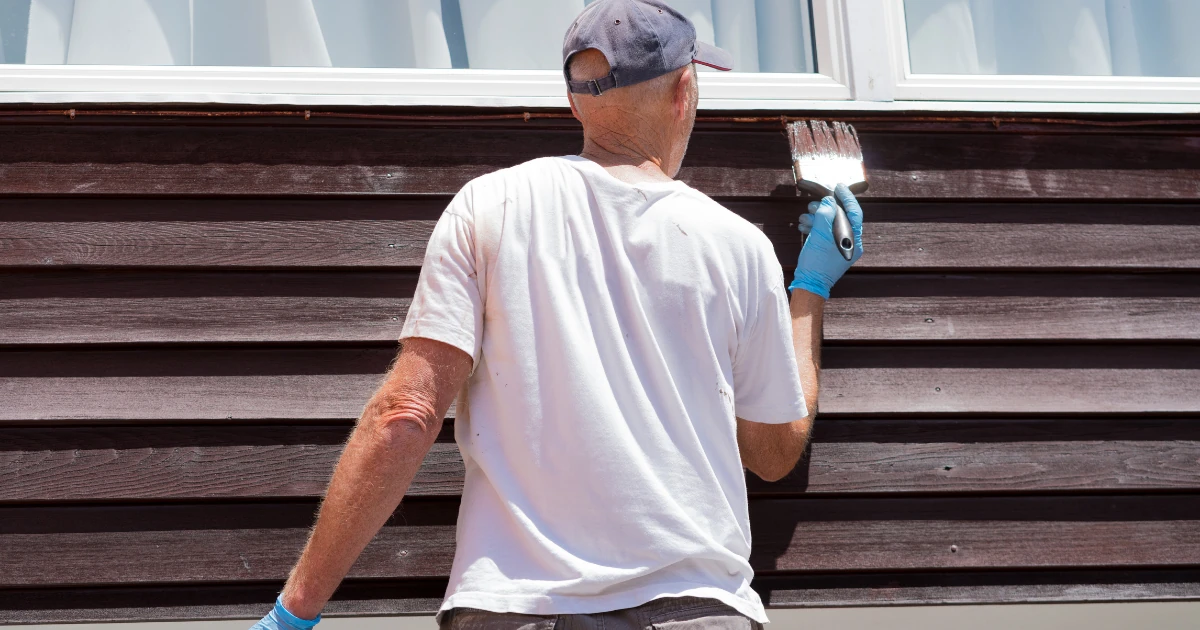 A man seen from behind painting dark wood siding trim near a window with a brush, wearing blue gloves and a white shirt.