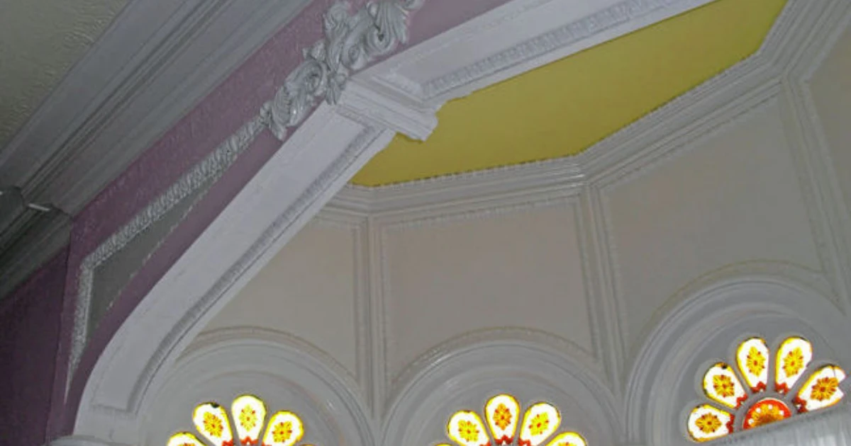 Upward view of ornate white crown molding and cornice work meeting purple and yellow painted wall and ceiling sections, above three stained-glass fan windows.