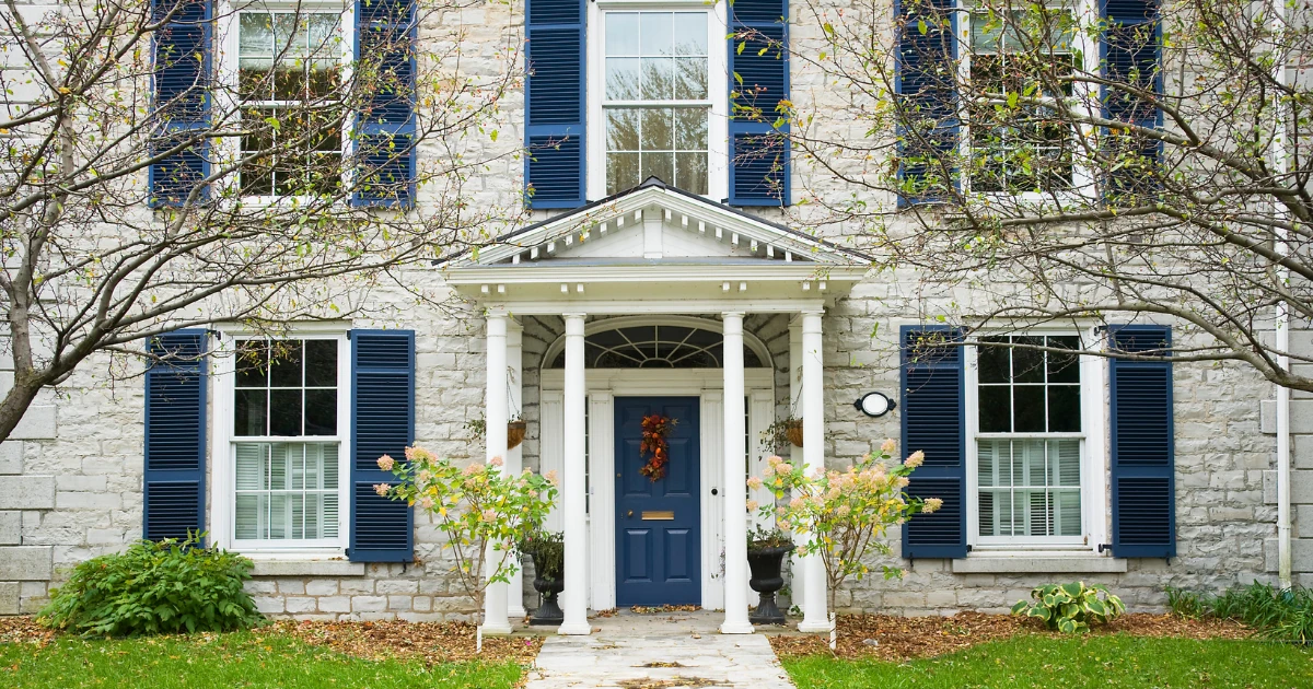 A stone house facade with navy blue shutters, white trim, a white columned portico, and a navy front door decorated with a fall wreath.