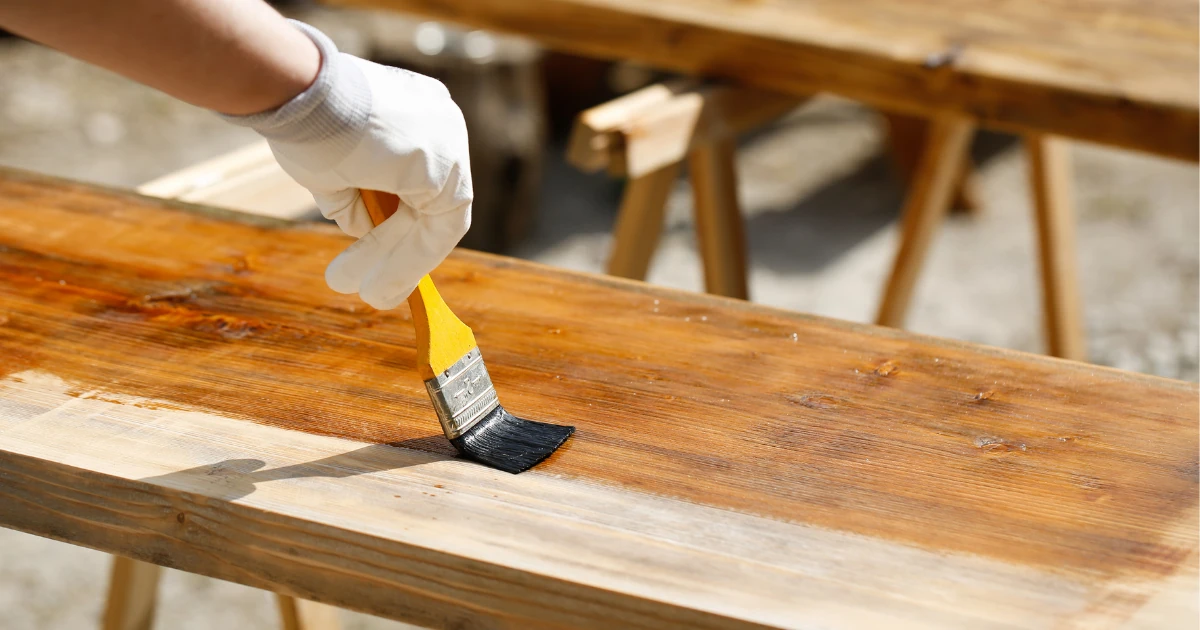 A gloved hand uses a paintbrush to apply stain or primer to a wood plank resting on sawhorses outdoors.