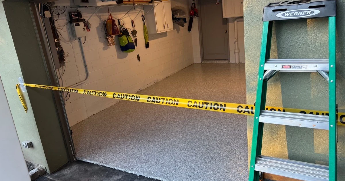 A garage doorway blocked by yellow caution tape, with a green ladder and a wet gray speckled epoxy floor coating visible inside.