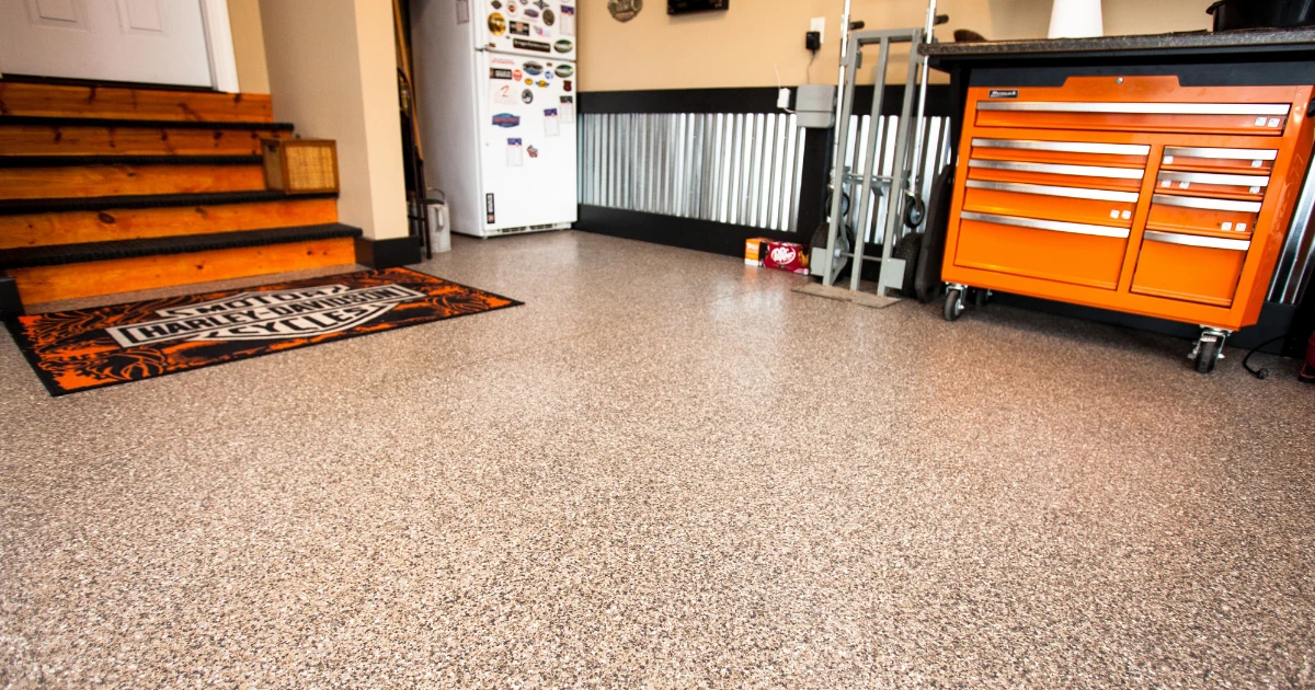 Garage floor with tan speckled epoxy coating, a Harley-Davidson floor mat near wooden stairs, a refrigerator, and an orange rolling tool chest.