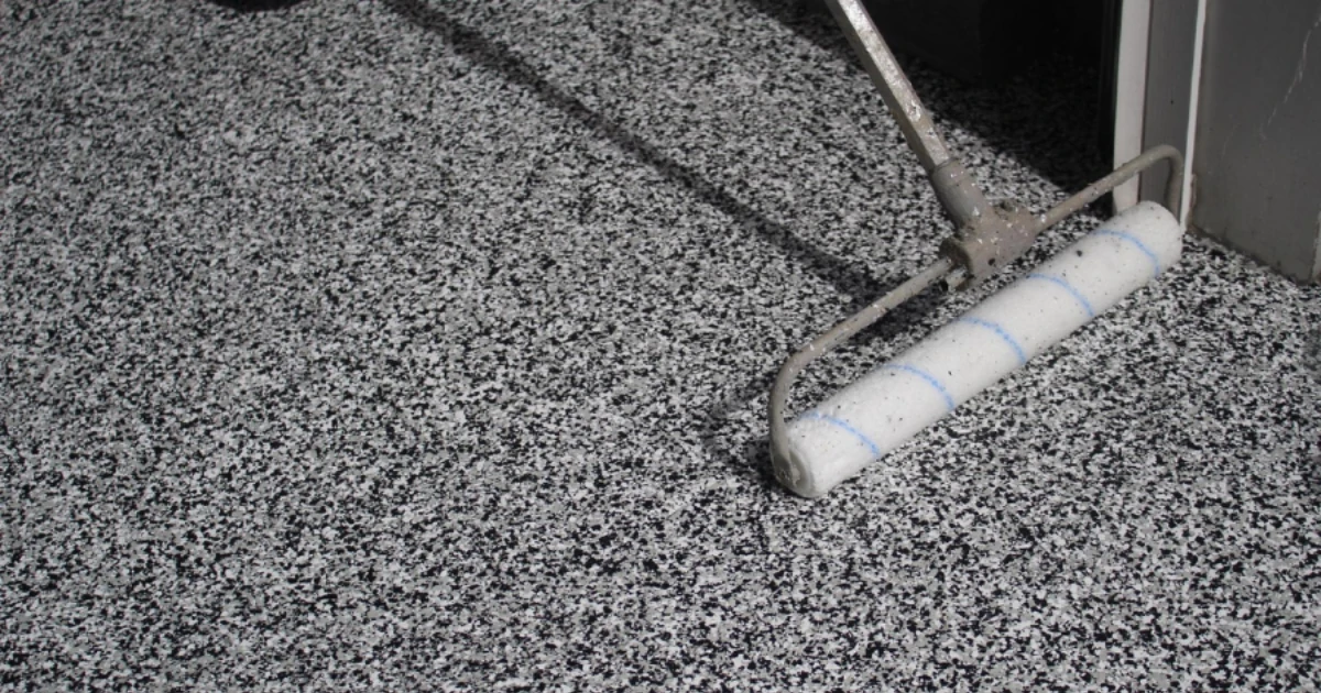 A paint roller resting on a gray speckled epoxy flake floor coating next to a doorway, with roller marks visible on the floor.