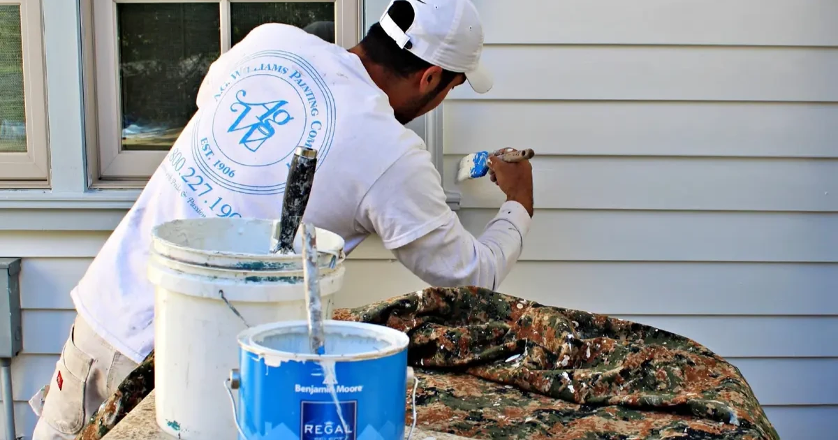 An A.G. Williams painter brushes light paint onto horizontal siding beside paint buckets.
