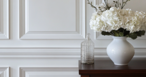 A close-up of a white, decorative wainscoted wall with intricate panel molding, complemented by a wooden table holding a white vase filled with white hydrangeas and a textured glass bottle.