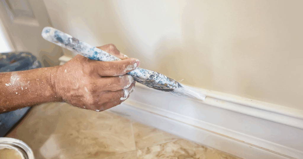 A close-up of a painter’s hand applying white paint to a baseboard with a precision brush, against a light beige wall.