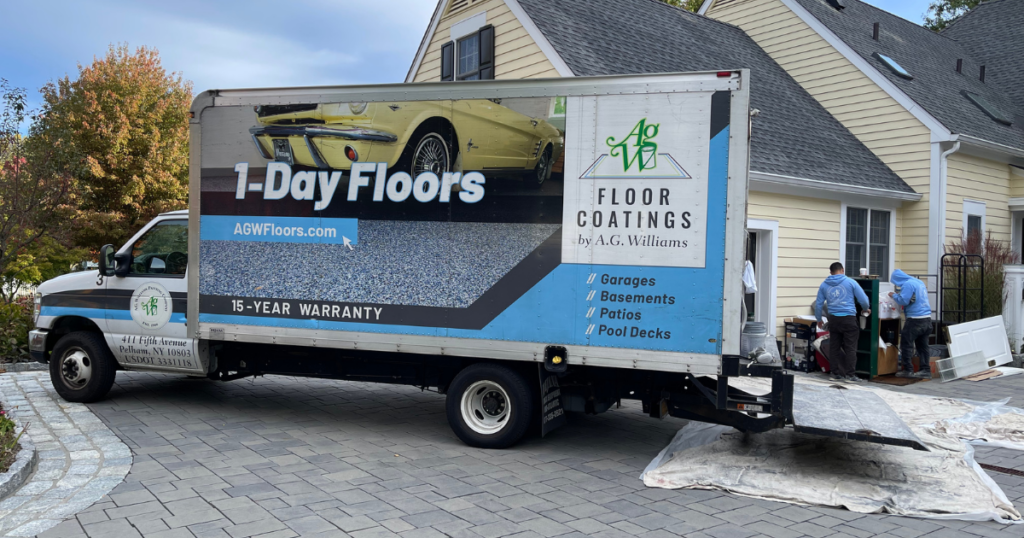 A branded A.G. Williams truck advertising 1-Day Floors and floor coating services is parked in the driveway of a residential home. Workers in matching uniforms are unloading equipment near the garage of a yellow house with a landscaped front yard.