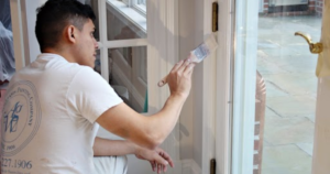 A professional painter carefully applies white paint to a door frame using a brush. The painter wears a white uniform, working with precision near a glass pane to achieve a clean and flawless finish.