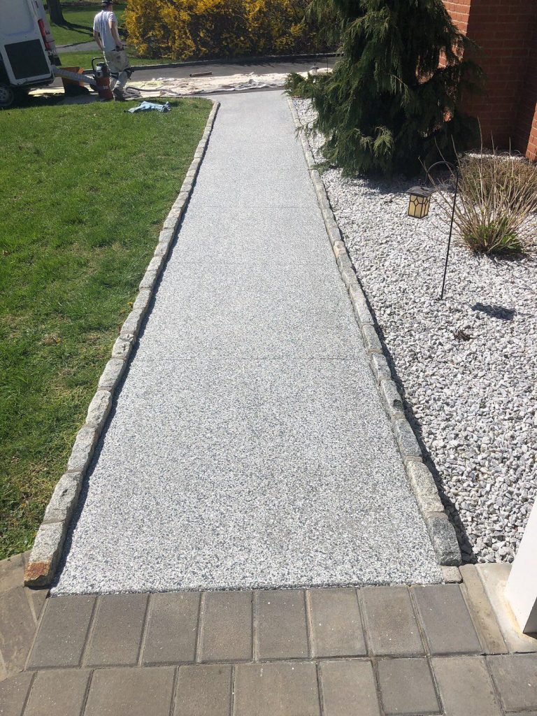 Outdoor walkway. Grey walkway that bumps up to brick. There is a grass edge on the left