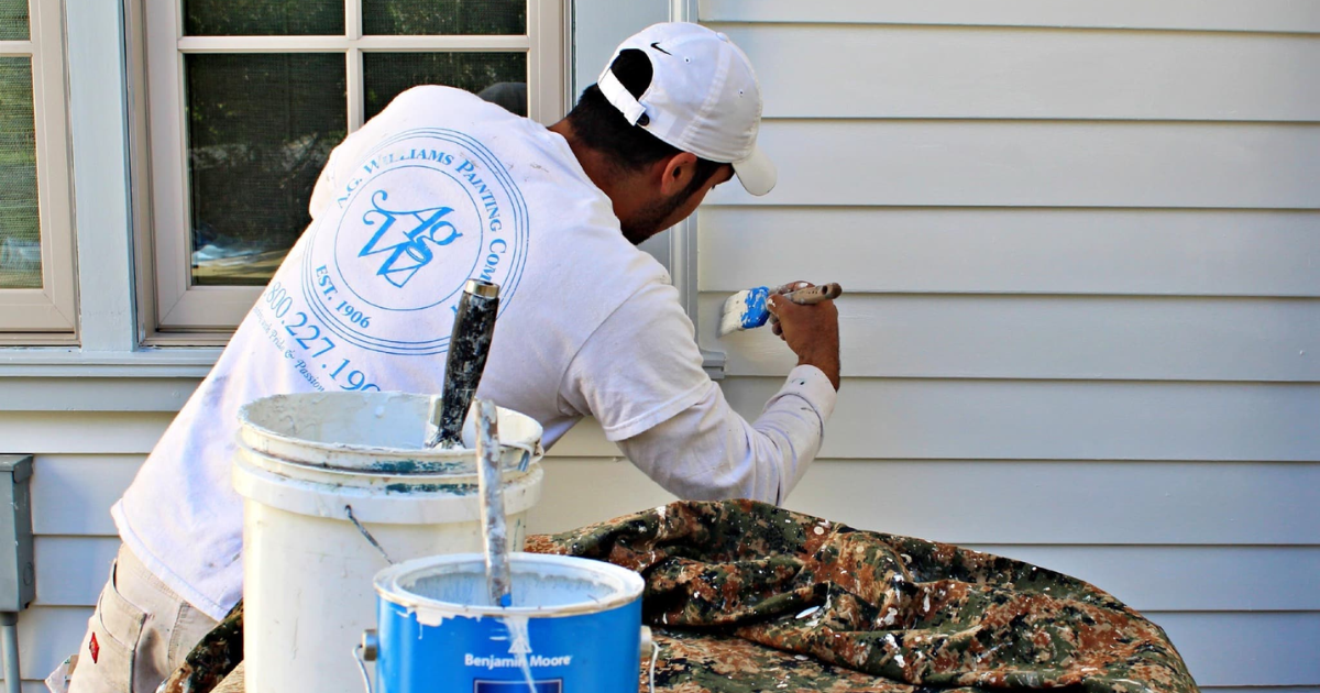 A professional painter in an A.G. Williams Painting Company uniform applies white paint to the exterior trim of a window, with buckets of paint and tools nearby, ensuring smooth and even application.