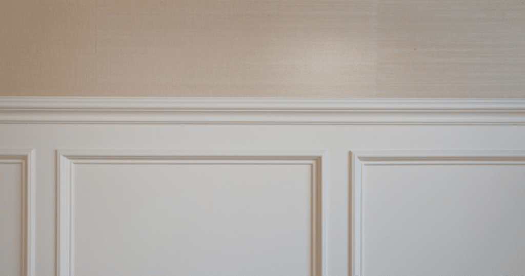 A close-up of a wall featuring a beige grasscloth wallpaper above white wainscoting panels with detailed molding. The combination adds texture and elegance to the space.