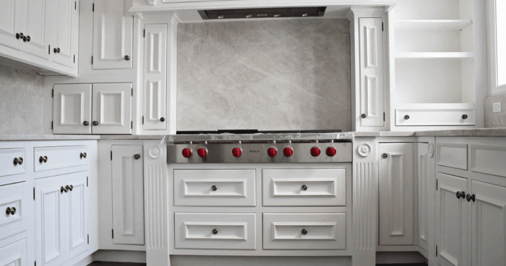 White kitchen with detailed cabinetry, marble backsplash, and a professional-grade stove with red knobs.