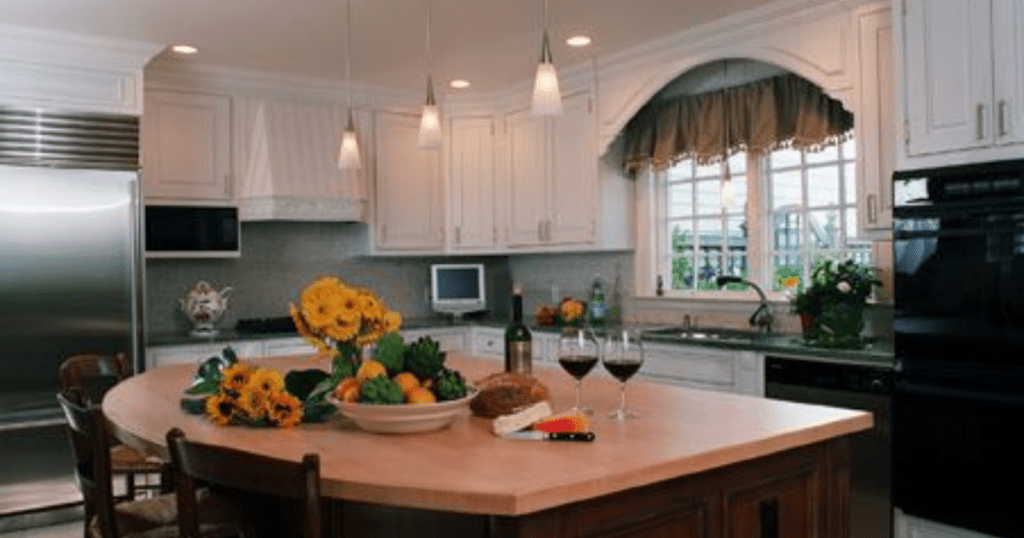 Staged kitchen with white cabinets, large windows, and a central island adorned with flowers, wine, and fresh produce.