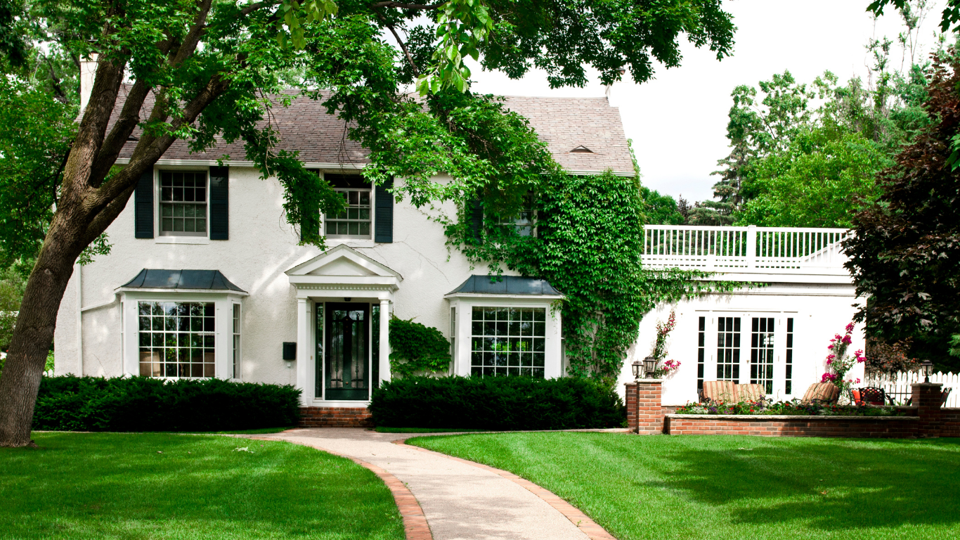 A classic ivy-covered colonial home with white stucco walls, black shutters, and a manicured lawn, surrounded by trees and colorful landscaping.