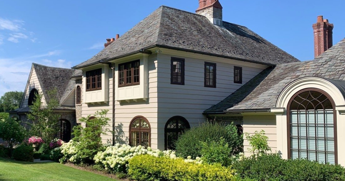 A beautifully maintained home with elegant cream-colored siding, brown-trimmed windows, and a slate roof, surrounded by vibrant landscaping with hydrangeas, manicured shrubs, and lush greenery under a bright blue sky.