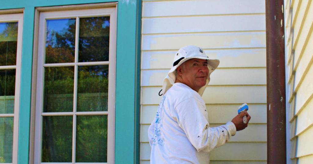 A professional painter working on the exterior of a house, wearing a sunhat and white painting attire, with a partially painted wall behind him.