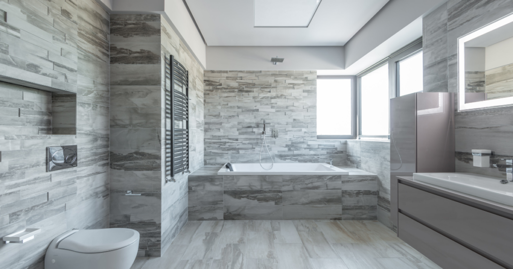 A contemporary bathroom with gray stone tile walls, a built-in bathtub, painted gray walls, and large windows allowing natural light.