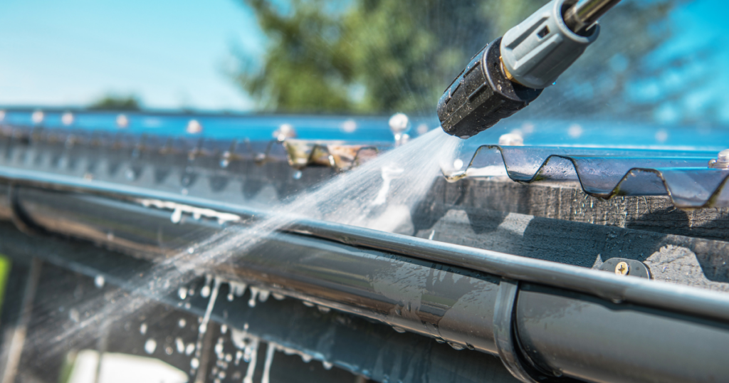 Close-up of a pressure washer cleaning a metal gutter, removing dirt and debris on a sunny day.