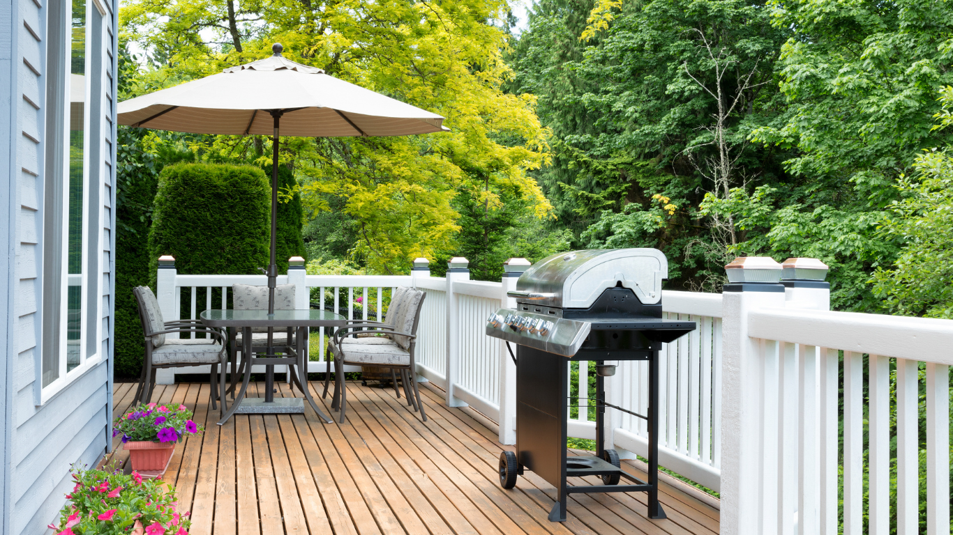 A wooden deck in Pleasantville, NY featuring a barbecue grill, patio furniture with a beige umbrella, and vibrant green trees in the background.