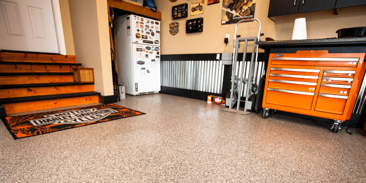 Garage with speckled epoxy floor, wooden stairs, refrigerator covered in magnets, orange tool chest, and Harley-Davidson mat, with motorcycle decor on the walls.