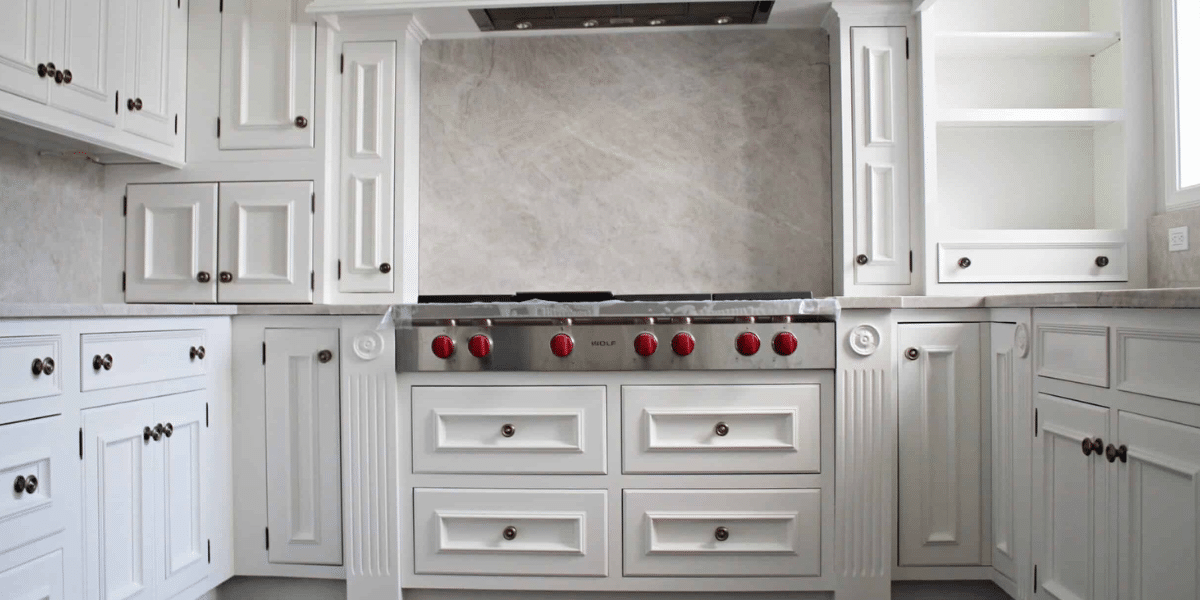 White kitchen cabinets with raised panel doors, bronze knobs, and a Wolf gas stove with red knobs. Light gray marble backsplash and open shelving on the right.