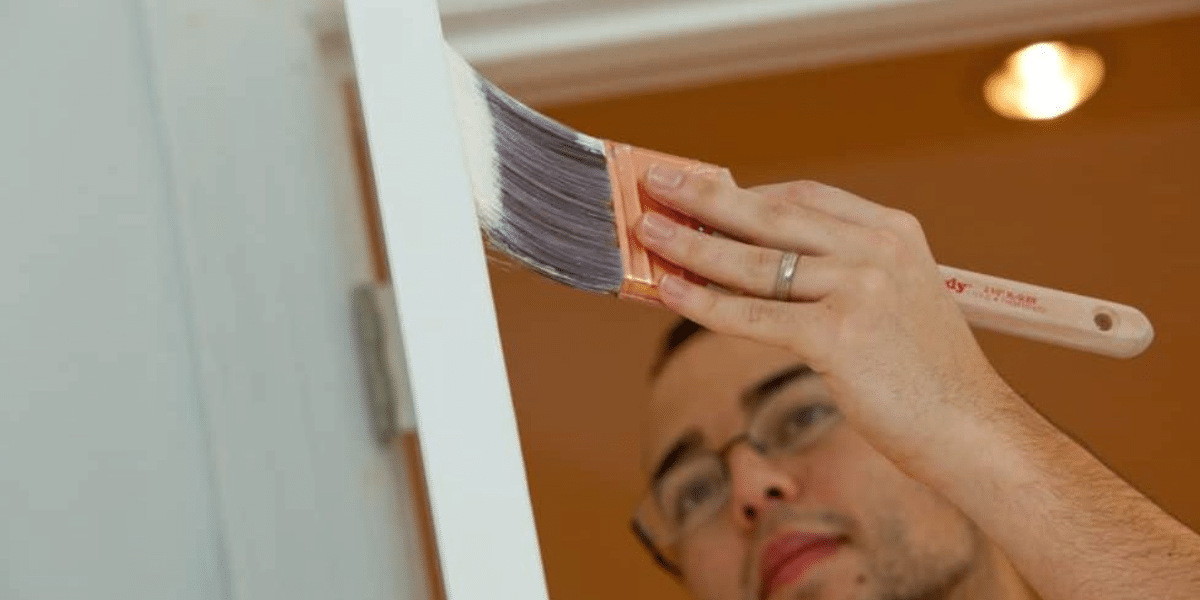 Person painting a door frame with white paint using a brush, focusing on precise detail work. The painter wears glasses and holds the brush at an angle for smooth application.