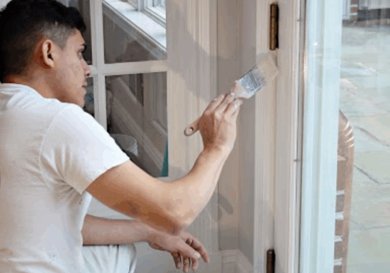 A professional painter carefully applies white paint to a door frame using a brush. The painter wears a white uniform, working with precision near a glass pane to achieve a clean and flawless finish.